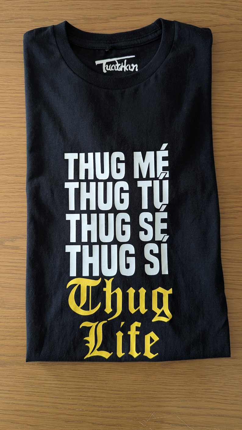 K&ouml;nnte beinhalten: Ein schwarzes T-Shirt mit kurzen &Auml;rmeln und wei&szlig;em Text, der THUG ME, THUG TU, THUG SE, THUG SI lautet, gefolgt von dem gelben Text THUG LIFE in einer gotischen Schriftart. Der Innenkragen zeigt das wei&szlig;e Markenlogo Tucutkun auf schwarzem Grund.