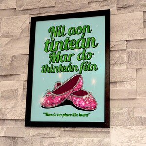 K&ouml;nnte beinhalten: Ein gerahmter Druck mit hellblauem Hintergrund und gr&uuml;nem Text, der "Nil aon tintean mar do tintean fein" und "There's no place like home" lautet. Der Druck zeigt ein Paar rosa glitzernde Schuhe.