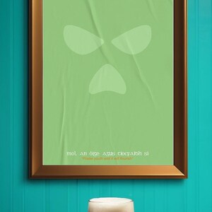Peut inclure: Une affiche verte encadr&eacute;e avec un design de visage stylis&eacute; et du texte en ga&eacute;lique, au-dessus d'une pinte de stout fonc&eacute;. L'affiche est dans un cadre marron, sur un mur turquoise. La stout a une t&ecirc;te &eacute;paisse et cr&eacute;meuse.
