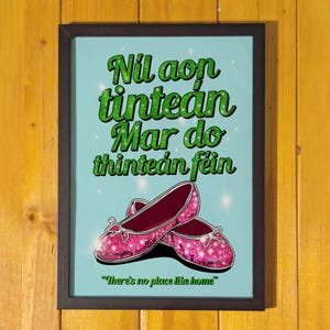 Puede incluir: Una impresión enmarcada con fondo azul y texto verde en gaélico irlandés que dice "Nil aon tintean mar do thintean fein." El texto está rodeado de zapatos rosas con purpurina. La parte inferior de la impresión dice "No hay lugar como el hogar."