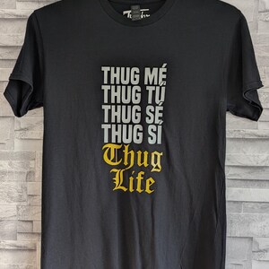 K&ouml;nnte beinhalten: Ein schwarzes Kurzarm-T-Shirt mit einem Grafikdruck auf der Brust. Der Text lautet THUG M&Eacute;, THUG TỰ, THUG S&Eacute;, THUG SI in grauen Gro&szlig;buchstaben, gefolgt von den Worten Thug Life in einer gelben gotischen Schrift unter dem grauen Text.