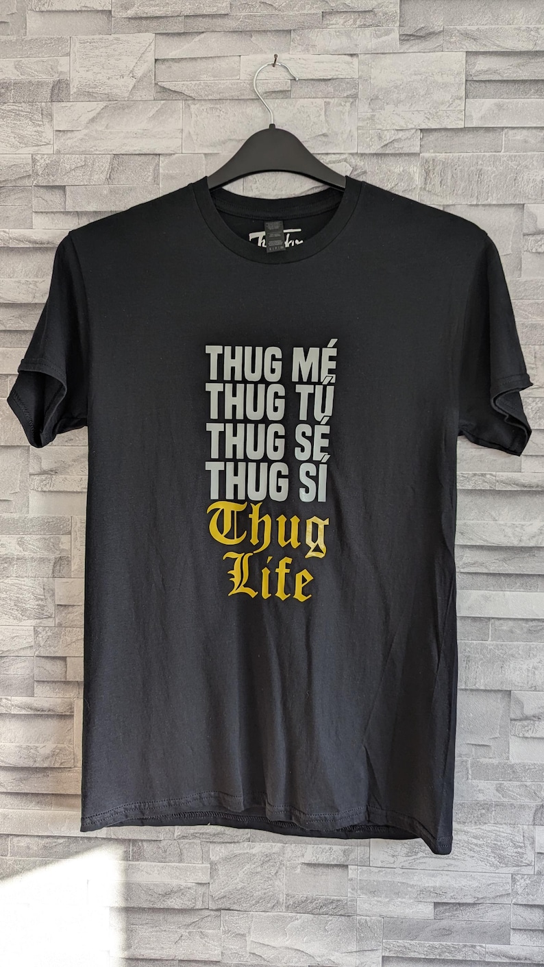K&ouml;nnte beinhalten: Ein schwarzes Kurzarm-T-Shirt mit einem Grafikdruck auf der Brust. Der Text lautet THUG M&Eacute;, THUG TỰ, THUG S&Eacute;, THUG SI in grauen Gro&szlig;buchstaben, gefolgt von den Worten Thug Life in einer gelben gotischen Schrift unter dem grauen Text.