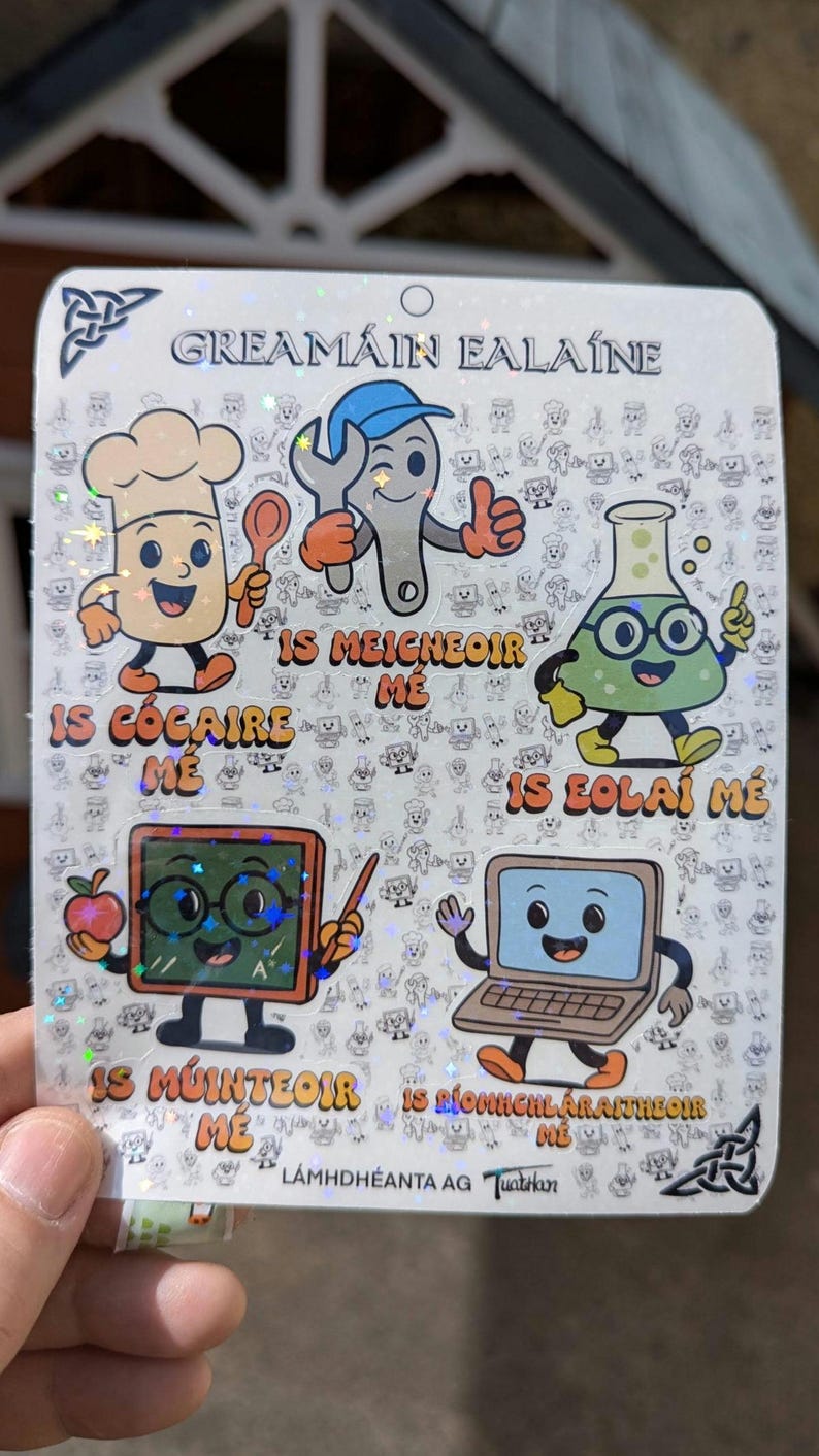 Peut inclure: Une feuille d'autocollants holographiques avec des personnages de dessins anim&eacute;s repr&eacute;sentant diverses professions. Les personnages incluent un chef, un m&eacute;canicien, un scientifique, un enseignant et un ordinateur. Du texte ga&eacute;lique est &eacute;galement pr&eacute;sent.