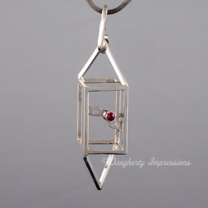 Puede incluir: Un collar con un colgante de plata de diseño geométrico que presenta una pequeña piedra preciosa roja. El colgante tiene forma de casa con un techo puntiagudo y una base cuadrada. La piedra preciosa está engastada en el centro del colgante.