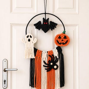 May include: A macrame wall hanging featuring a black circular hoop with a central black bat, a white ghost, and an orange pumpkin. Below, vertical strands of black, orange, and white cord are arranged with a black spider attached to the center.
