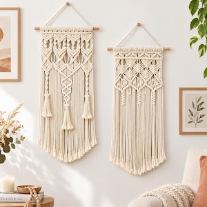 May include: Two off-white macrame wall hangings featuring intricate knotted patterns, suspended from horizontal wooden dowels with attached hanging cords. The designs include geometric diamond shapes and long, loose fringe at the bottom edges.