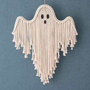 May include: A wall hanging crafted from off-white cotton cord, shaped like a ghost with a rounded head and outstretched arms. The design features two oval black felt eyes and a circular black felt mouth, with long, loose fringe hanging down.