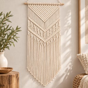 Peut inclure: Une tenture murale en macramé de couleur crème présentant des motifs de nœuds complexes en forme de chevron, suspendue à une tige en bois horizontale. La pièce se compose de plusieurs couches de cordes tressées finissant en longues franges.