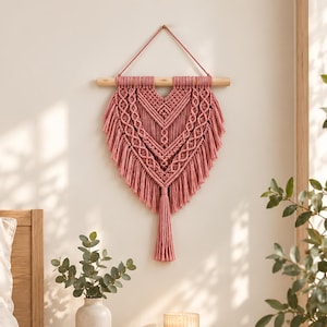 May include: A heart-shaped macrame wall hanging crafted from dusty rose cord. The piece features intricate knotted patterns, a central V-shaped design, and long fringe ending in a single tassel, all suspended from a horizontal wooden dowel.