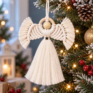 May include: A macrame angel ornament crafted from off-white cotton cord, featuring a smooth wooden bead head, a circular wooden halo, fringed wings, and a long tassel skirt accented with a small black star at the waist, suspended by a cord loop.