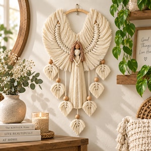 May include: A macrame wall hanging featuring a central figure with brown hair and a cream-colored cord body, flanked by large, wing-like cord structures. The piece includes several leaf-shaped macrame accents and round wooden beads.