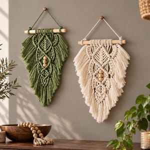 May include: Two macrame wall hangings suspended from wooden dowels. One is olive green and the other is off-white. Both feature intricate knotted patterns, fringed edges, and three central wooden beads. Each is hung by a cord from a wall hook.