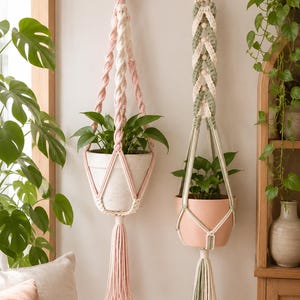 May include: Two macrame plant hangers suspended from wooden wall hooks. The left hanger features pink and cream twisted cord with a pink tassel. The right hanger uses sage green and cream braided cord with wooden beads and a sage green tassel.