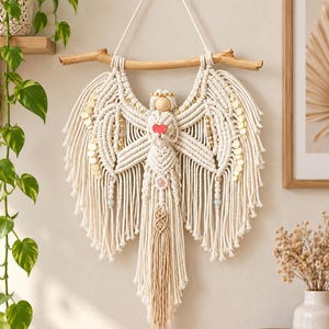 May include: A macrame angel wall hanging crafted from off-white cotton cord, suspended from a natural wooden branch. The figure features a round wooden head, expansive fringed wings accented with gold sequins, and a small red heart.