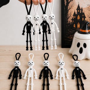 May include: A collection of macrame skeleton ornaments featuring white wooden bead heads with black painted facial features, including x-shaped eyes and a stitched mouth. The bodies are crafted from black or white cord with knotted limbs.
