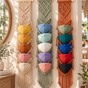 May include: Three vertical macrame wall organisers made of cotton cord, each featuring five integrated pockets holding baseball caps in various colours. The left organiser is cream, the centre is sage green, and the right is burnt orange.