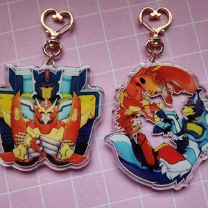 May include: Two clear acrylic keychains with gold heart-shaped rings. The keychain on the left features a red, yellow, and blue robot character. The keychain on the right features a red, blue, and white furry character.
