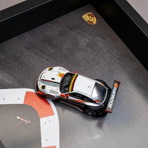 Può includere: Modello incorniciato di un'auto da corsa Porsche 911 GT3 RS. L'auto è bianca, nera e arancione, con loghi degli sponsor. L'opera d'arte include un design di pista e l'emblema Porsche. La cornice è nera.