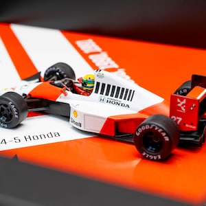 Peut inclure: Maquette détaillée d'une voiture de course McLaren MP4-5 Honda de Formule 1, blanche et orange. Le modèle présente des pneus noirs avec la marque Goodyear, un logo Shell et le numéro 27. La voiture est posée sur un fond orange et blanc vif.