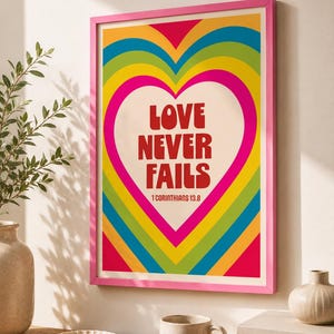May include: A rectangular wall art print in a bright pink frame featuring a large central heart shape surrounded by concentric rainbow-coloured arches in red, orange, yellow, green, and blue. The text reads LOVE NEVER FAILS 1 CORINTHIANS 13.8.