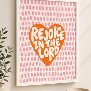 May include: A framed wall art print featuring a large orange heart centred on a light pink background with a pattern of small, vertical, dashed brushstrokes. The text "REJOICE IN THE LORD" is written in bold, white, rounded capital letters.
