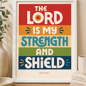 May include: A white framed wall art print featuring the text THE LORD IS MY STRENGTH AND SHIELD and PSALM 28.7. The design uses horizontal colour blocks in red, orange, yellow, green, and dark blue with bold, cream-coloured sans-serif typography.