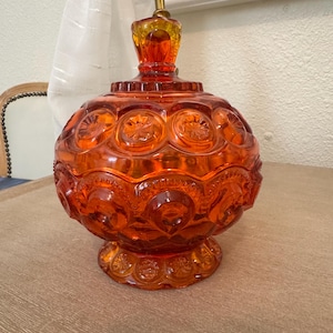 May include: A round, orange glass candy dish with a lid, featuring a raised star and moon pattern across its surface. The dish sits on a pedestal base and includes a matching glass lid with a knob handle, showcasing a vibrant, translucent finish.
