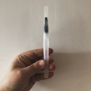 Op de afbeelding: Een transparante waterverf penseel pen van plastic met een zwarte punt. De pen wordt in een hand gehouden.