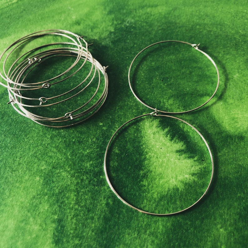 May include: A collection of silver-colored hoop earrings, some stacked and others separate, against a vibrant green watercolor background. The earrings are simple, round, and have a small clasp.