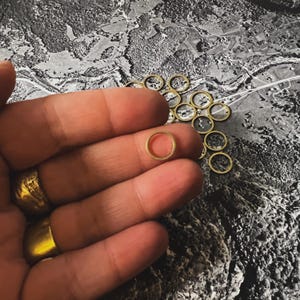 May include: A close-up of a hand holding a small, gold-colored metal ring. The ring is about 1 cm in diameter and has a smooth, polished finish. There are many other similar rings scattered on a textured surface in the background.