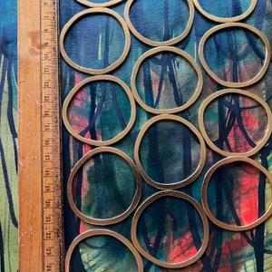 May include: A collection of ten gold-colored metal rings of varying sizes, arranged on a dark blue and red background with black brushstrokes. The rings are all circular and have a smooth, polished finish.