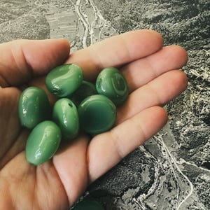 May include: A collection of smooth, oval-shaped, green beads. The beads have a glossy finish and vary slightly in size. Some beads have a hole drilled through them, suggesting they are for stringing. The beads are held in a hand against a black and white background.