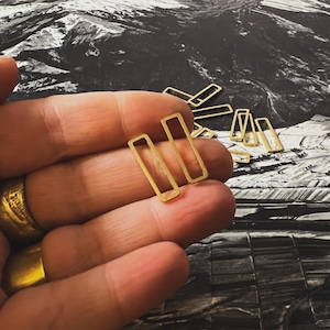 May include: Gold-colored rectangular earrings. The earrings are held in a hand, with several other similar earrings visible in the background. The earrings have a simple, minimalist design, with a rectangular shape and a hollow center.