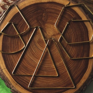 Puede incluir: Cuatro pendientes de aro geométricos de color dorado, incluyendo un triángulo, un hexágono y dos semicírculos, dispuestos sobre una superficie de madera.