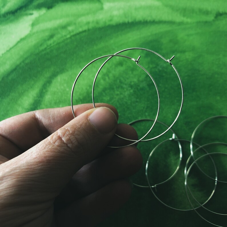 May include: A hand holding several silver-colored hoop earrings against a green background. The earrings are thin and circular, with a simple clasp design. The background has a watercolor-like texture.