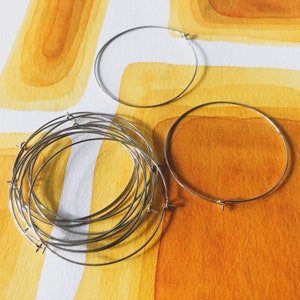 May include: A collection of silver-colored metal hoop earrings of varying sizes. Some hoops are stacked together, while others are displayed individually. The background features an abstract watercolor design in shades of yellow and orange.