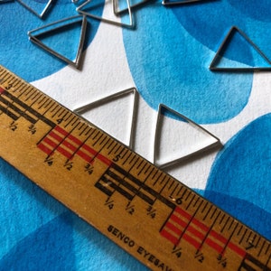 May include: A close-up of a wooden ruler with markings in inches and fractions of an inch. The ruler is lying on a white surface with a blue watercolor background. There are several silver triangle shapes scattered on the surface.