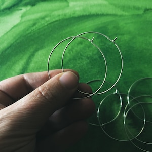 May include: A set of silver hoop earrings, with one hoop slightly larger than the other, held in a hand against a green watercolor background.