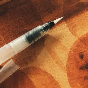 Op de afbeelding: Een transparante waterverf penseelpen van plastic met een zwarte punt en een witte borstel. De pen ligt op een bruin en beige, getextureerd oppervlak.