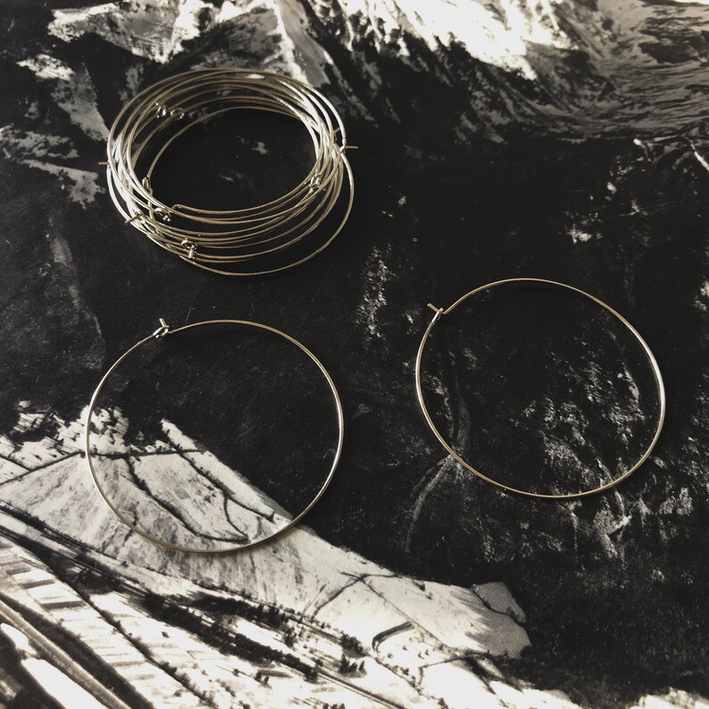 May include: A collection of silver-colored hoop earrings in various sizes, some stacked together, others laid out individually. The earrings are set against a black and white landscape background, creating a minimalist aesthetic. The hoops appear to be made of thin metal.