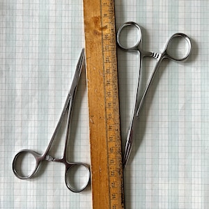 May include: Two surgical clamps, one with a straight handle and one with a curved handle, are laid out on a gridded surface next to a wooden ruler with markings from 7 to 25.