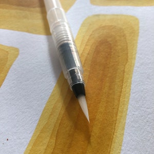 Op de afbeelding: Een witte aquarel penseelstift met een zwarte punt. De pen ligt op een vel papier met gele aquarelverf.