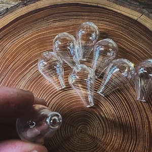 May include: Clear glass teardrop-shaped objects with a swirl pattern are arranged on a wooden surface. One is held by a hand. The glass has a slight iridescent quality, reflecting light. The objects appear to be small, possibly decorative or functional.