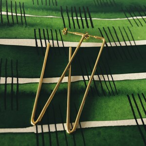 May include: A pair of gold-toned geometric earrings with a triangular shape. The earrings are on a green and white striped background.