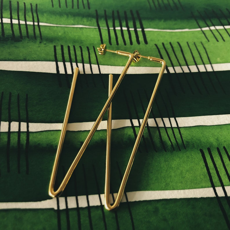May include: A pair of gold-toned geometric earrings with a triangular shape. The earrings are on a green and white striped background.