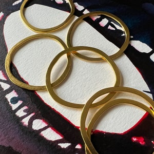 May include: Five gold-toned metal rings of varying sizes, stacked and overlapping, creating a circular pattern. The rings are arranged on a black and white background with red and white abstract patterns.