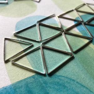 May include: A close-up of a silver geometric design made of triangles. The design is on a white background with green and blue watercolor washes.