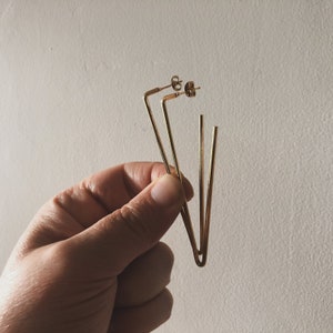 May include: A pair of gold-toned earrings with a geometric design. The earrings are made of thin metal rods that are bent into a triangular shape.