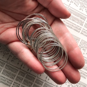 May include: A collection of silver-colored metal rings, some with a black finish, are held in a person's hand. The rings are thin and circular, and they are stacked together. The background is a printed page with numbers and text.