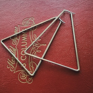 Puede incluir: Un par de pendientes de aro triangulares geométricos de color plateado. Los pendientes se muestran sobre una superficie roja con letras doradas. Los pendientes tienen un diseño simple y moderno, con forma triangular. Los pendientes son de metal brillante.